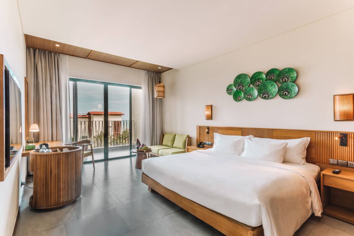 A hotel room with a large bed, a desk, a chair, a small table, a green wall decoration, and a view of the outside.