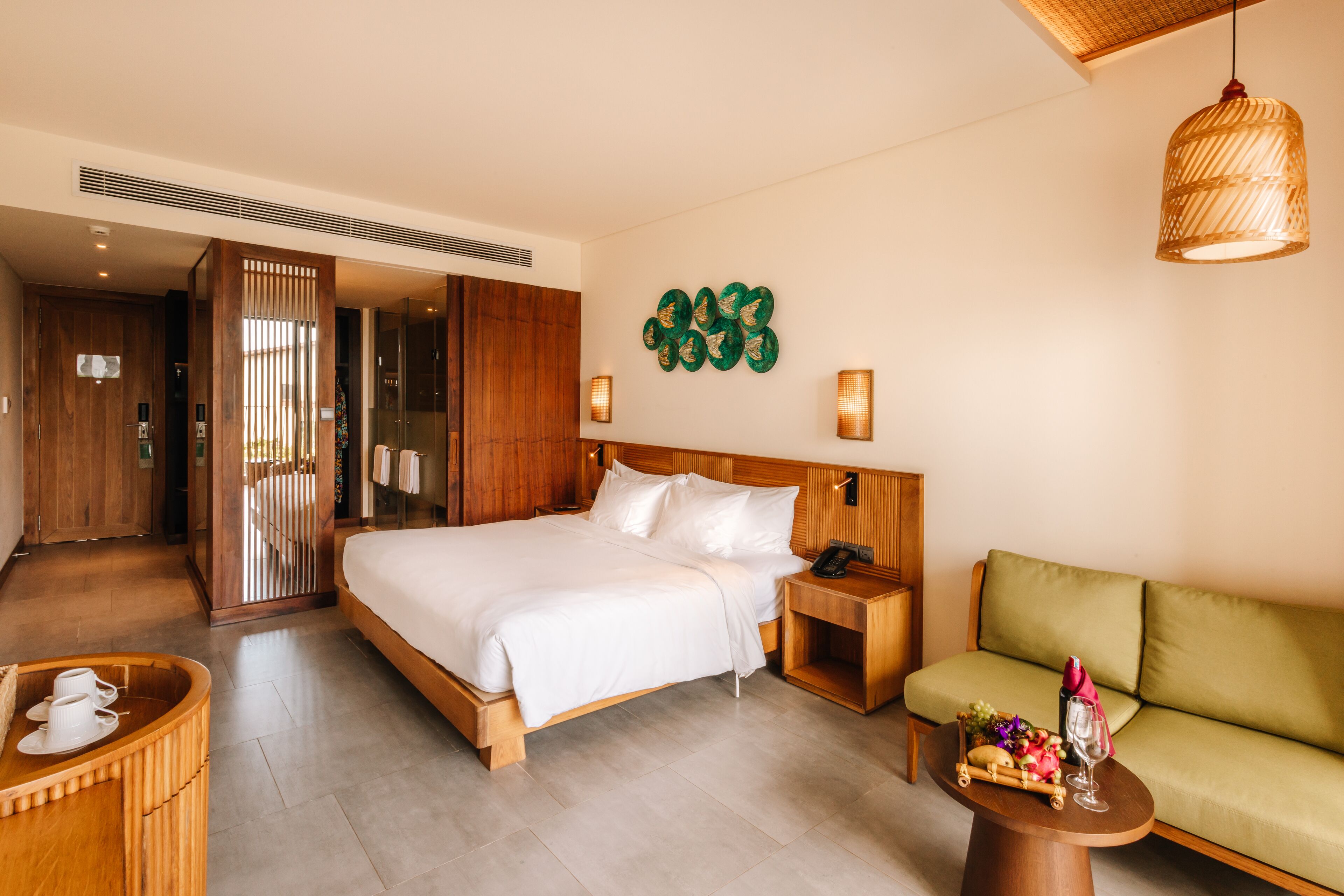 Una habitación de hotel moderna con una cama grande, cabecera de madera, un sofá verde, una pequeña mesa de madera con una cesta de frutas y una lámpara de pared.