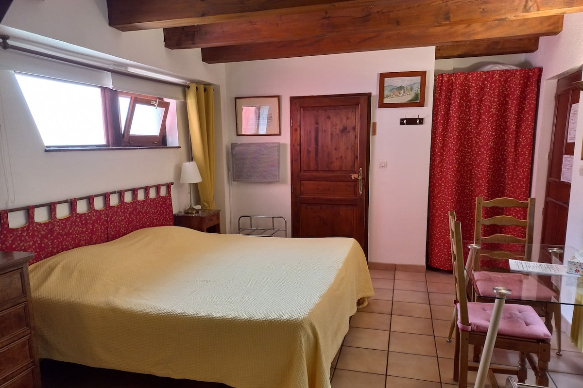 A bedroom with a bed, wooden headboard, a small table with a chair, a wooden door, and a window with curtains.