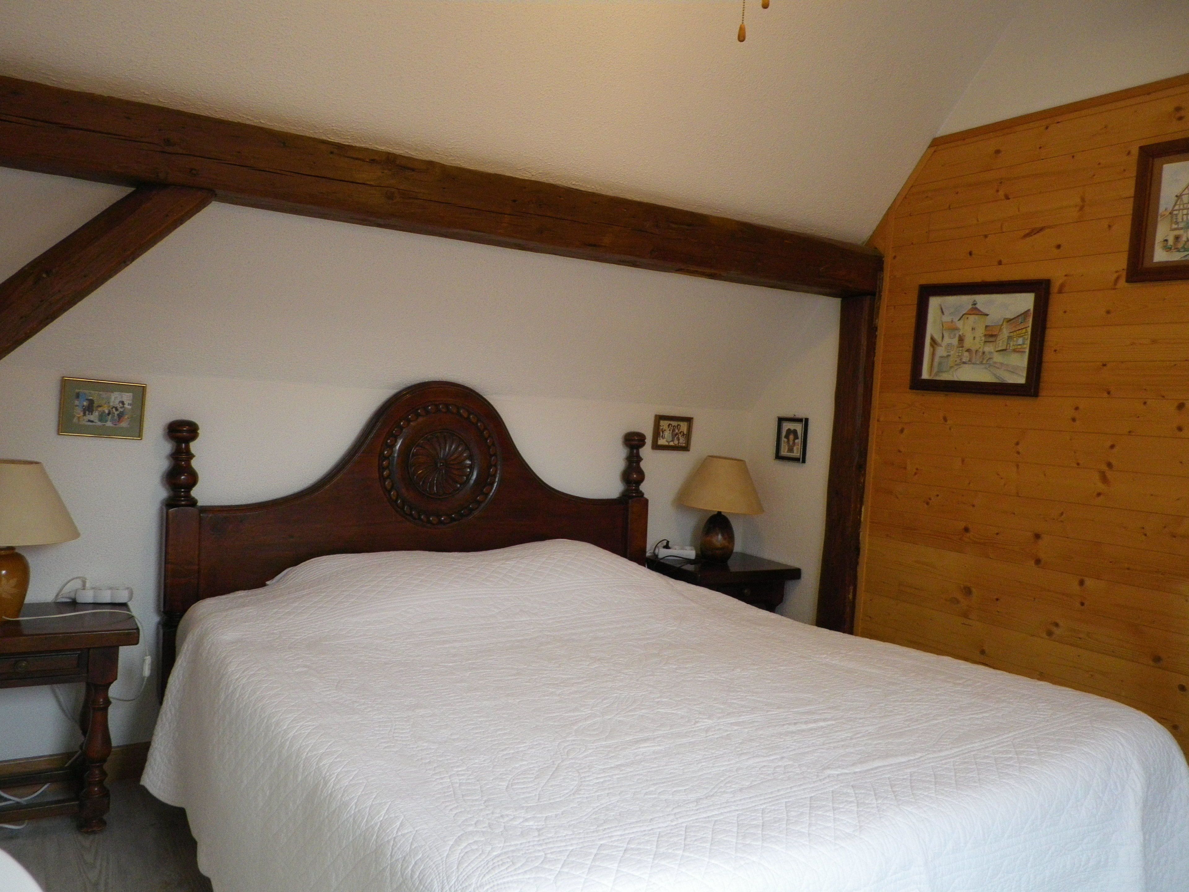 A bedroom with a wooden headboard, a bed with a white quilt, two bedside tables with lamps, and a wooden wall with framed pictures.