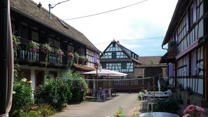Una estrecha calle adoquinada flanqueada por casas tradicionales con entramado de madera, mesas al aire libre y plantas con flores.