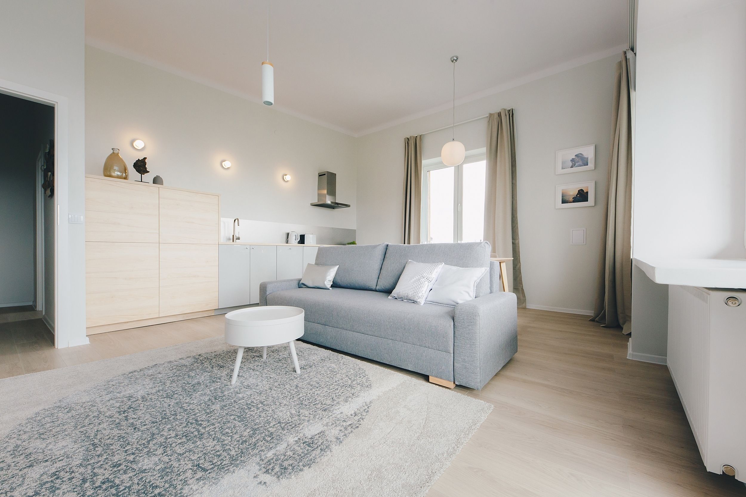 A modern living room with a grey sofa, a white coffee table, and a light-colored rug. There is a kitchen area with a white cabinet and a hanging light fixture.