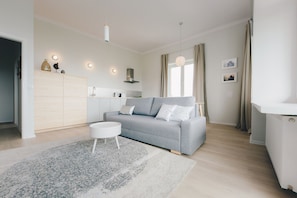 A modern living room with a grey sofa, a white coffee table, and a light-colored rug. There is a kitchen area with a white cabinet and a hanging light fixture. - ShortStayPoland Świętokrzyska - B38 (Warsaw)