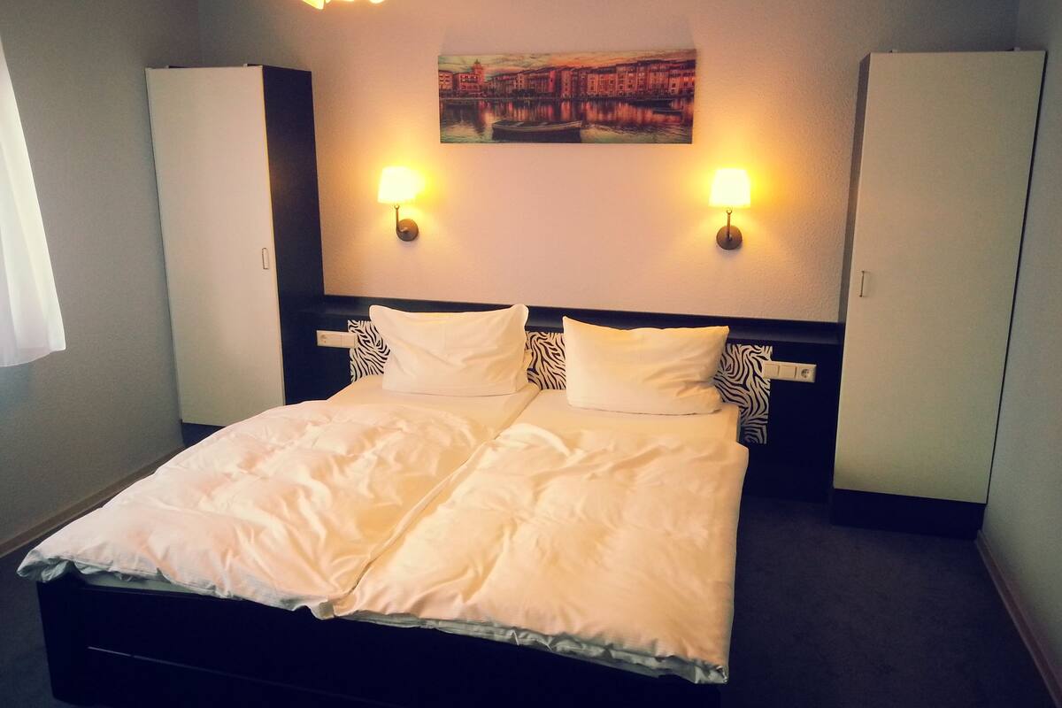 A hotel room with a large bed, two wall-mounted lamps, a painting above the bed, and a wardrobe on either side.