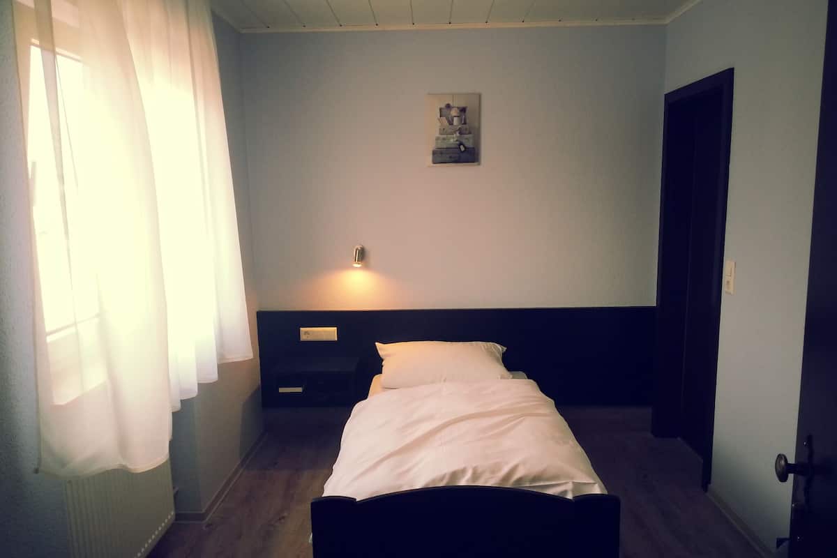 A small, simple room with a single bed, a nightstand, a wall-mounted light, and a door.