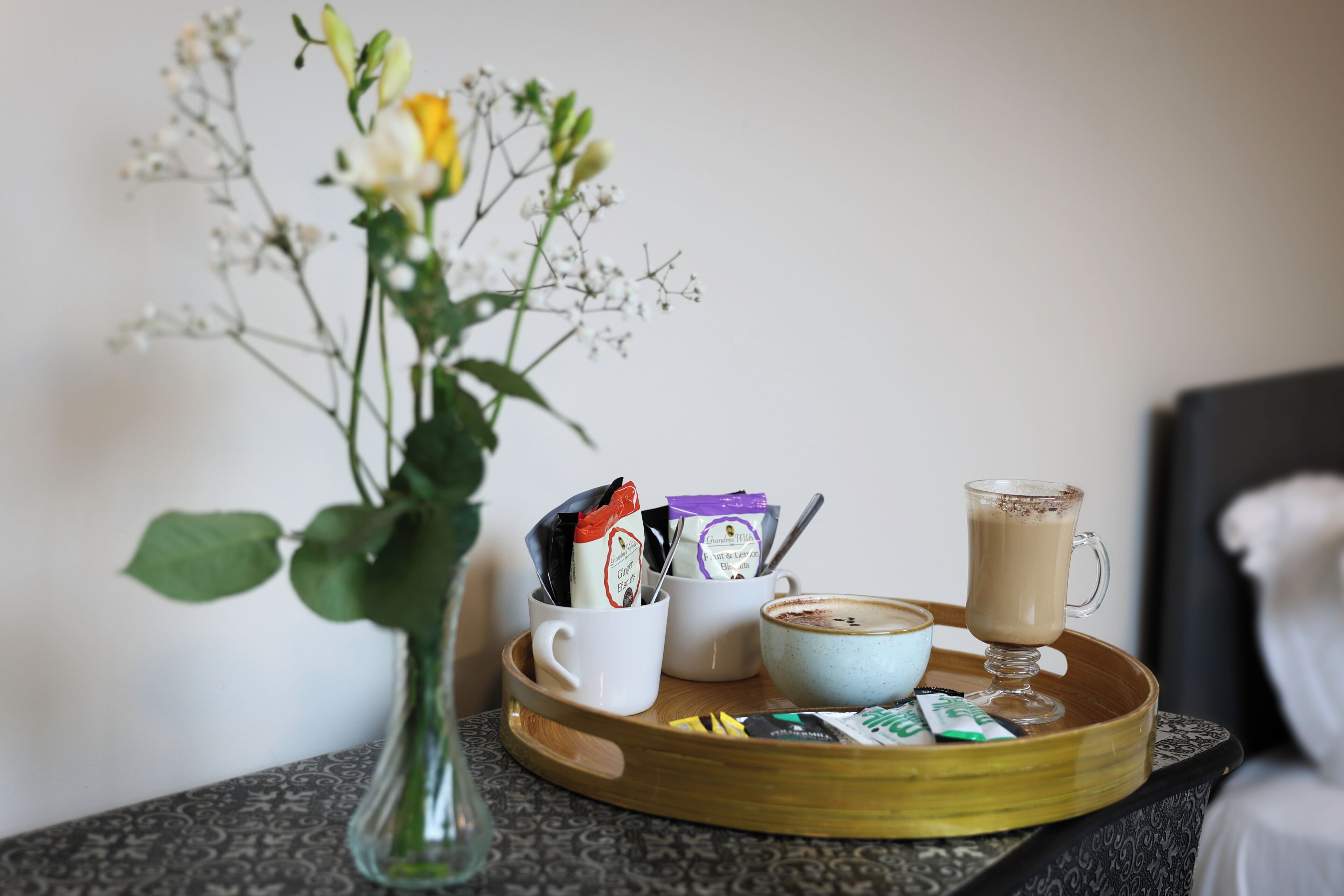 A tray with a coffee cup, a glass of coffee, a vase with flowers, and some packets on a table.