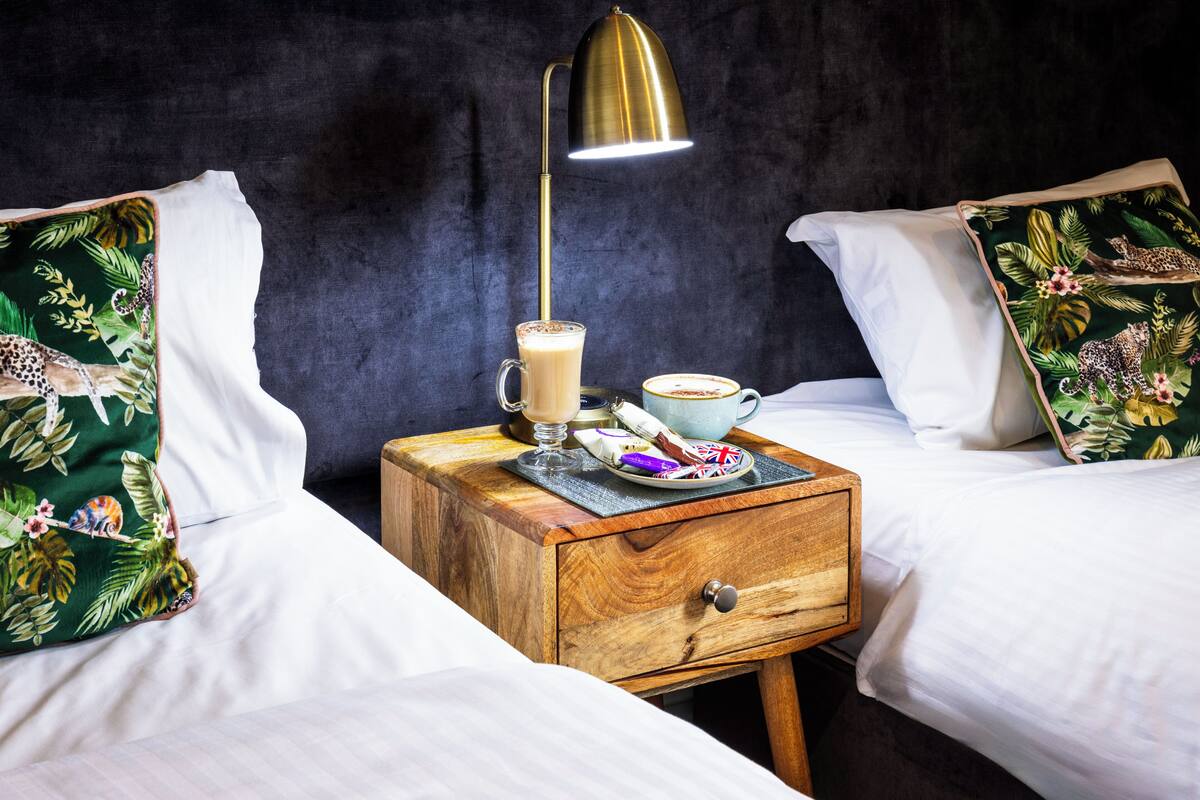 Quarto de hotel com duas camas, uma mesinha de cabeceira com abajur, uma xícara de café e um prato com lanches.