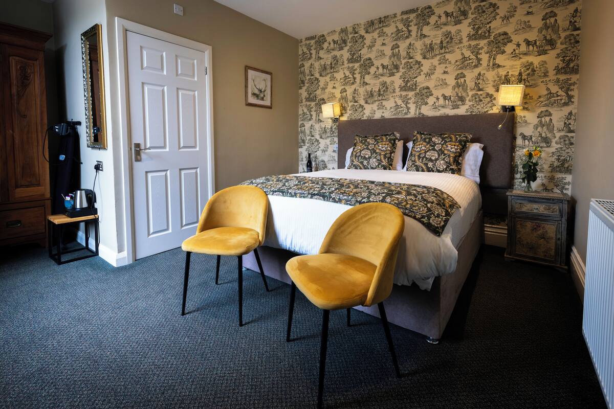 Quarto de hotel com cama, duas cadeiras, guarda-roupa, espelho, abajur e papel de parede estampado.