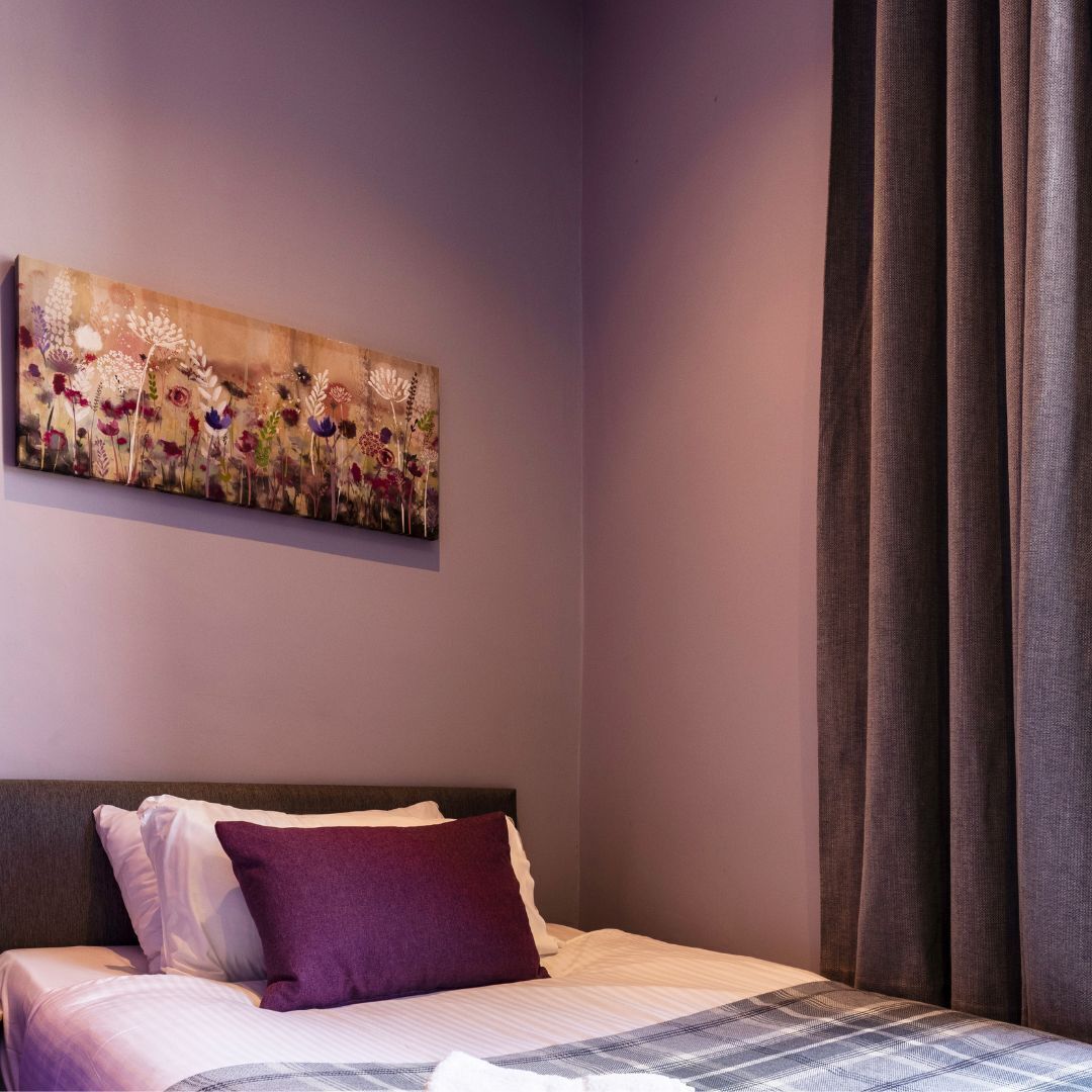A bedroom with a bed, a purple pillow, a painting on the wall, and a window with curtains.