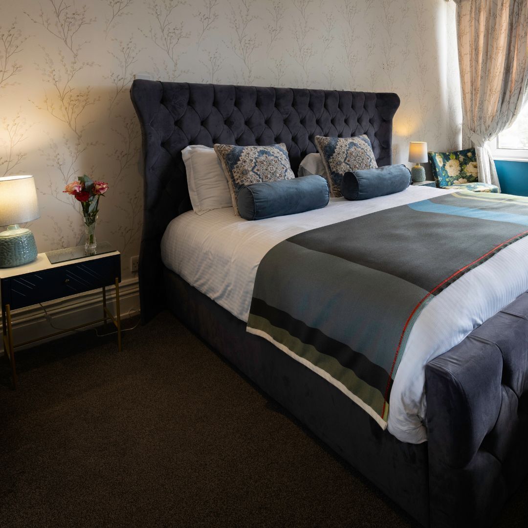 A bedroom with a tufted headboard, a bed with multiple pillows, a bedside table with a lamp, and a vase with flowers.