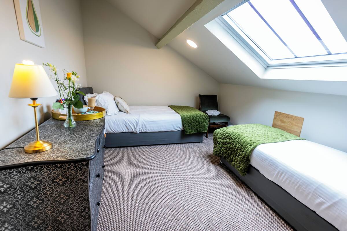 Quarto com claraboia, duas camas, mesa de cabeceira com abajur e um vaso com flores.