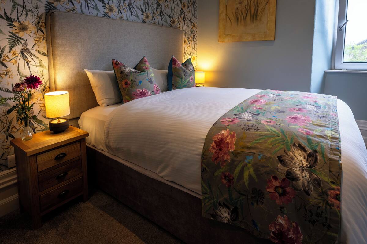 Uma cama bem arrumada com roupa de cama florida, uma luminária de cabeceira e uma mesa de cabeceira com gavetas.