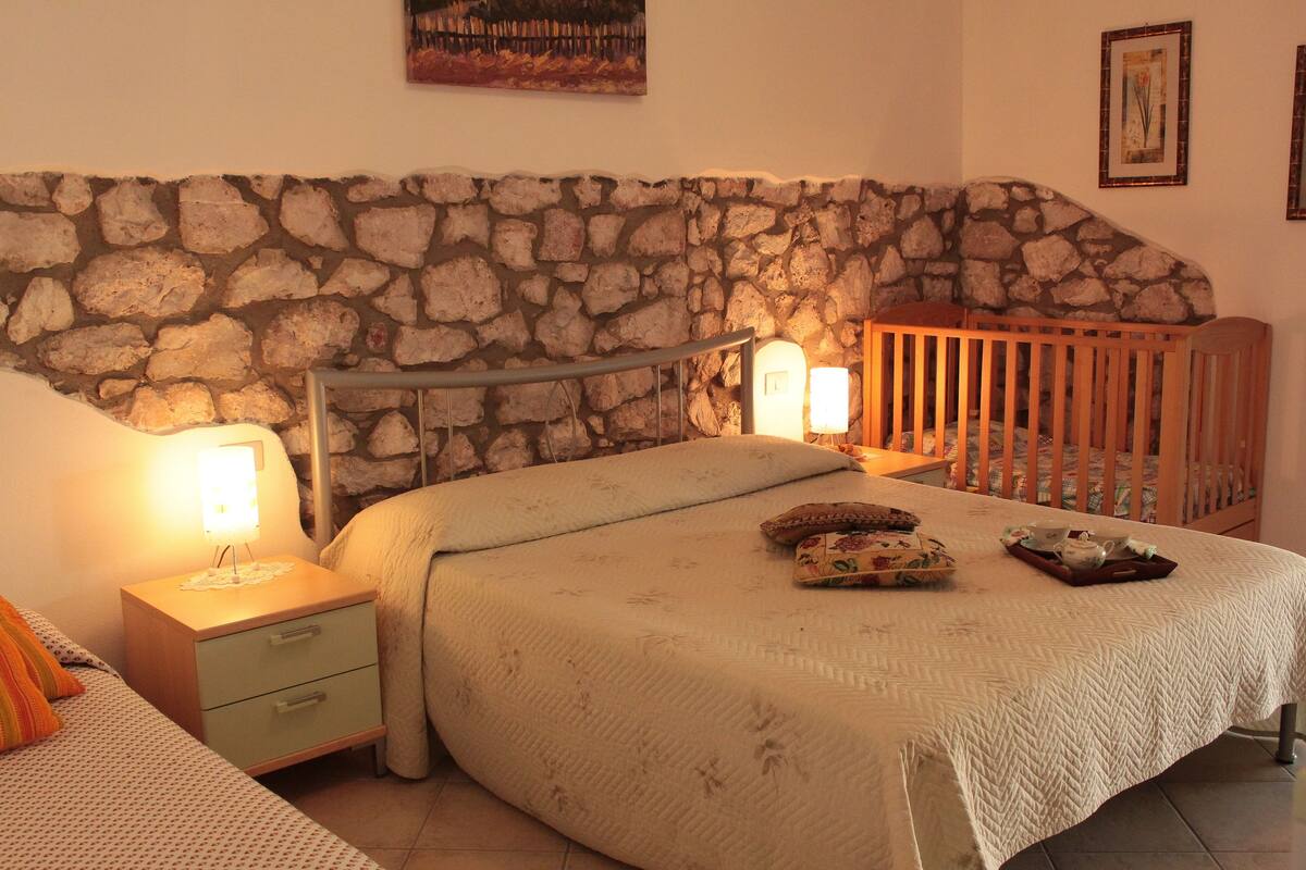 Un dormitorio con pared de piedra, una cama con colcha, una mesita de noche, una cuna de madera y un cuadro enmarcado en la pared.