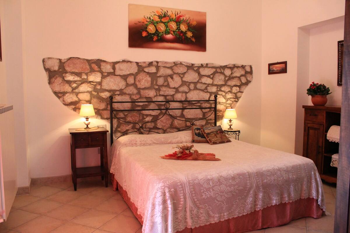 Un dormitorio con cabecera de piedra, dos mesitas de noche con lámparas, una cama con cubrecama de encaje y un cuadro floral sobre la cama.