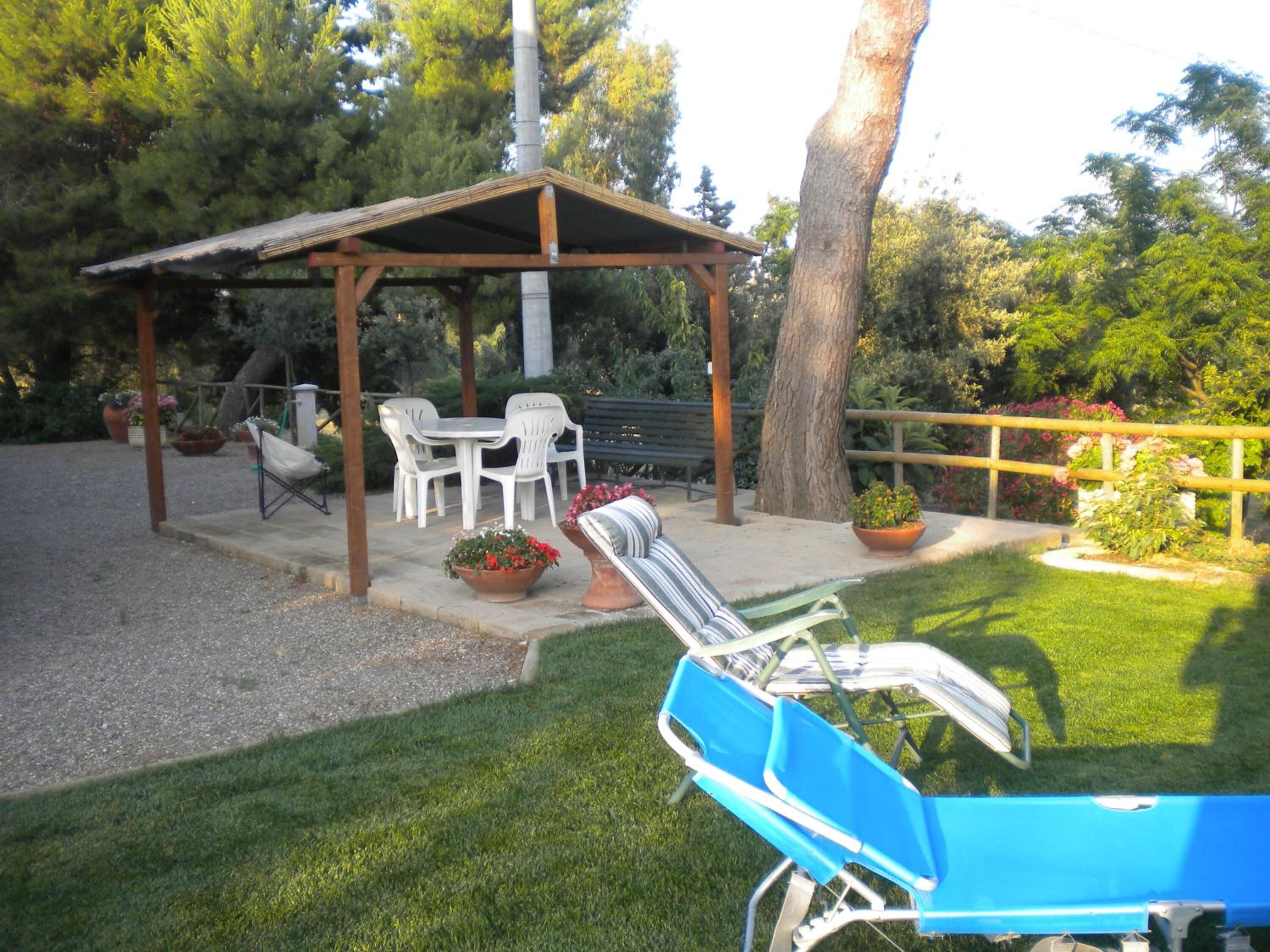 Un patio couvert, aménagé avec du mobilier d’extérieur, notamment des chaises et une table, et entouré d’une clôture en bois et d’une végétation luxuriante.