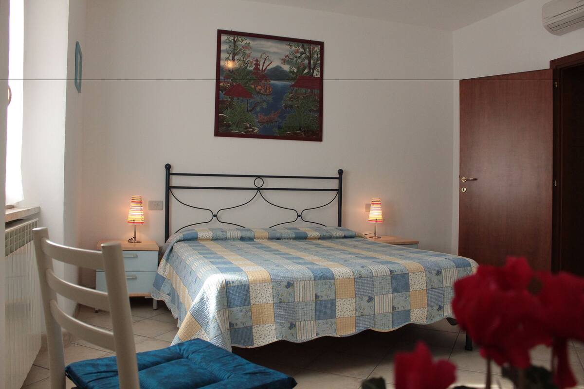 Un dormitorio con cama, lámparas de noche, una silla, una puerta de madera y un cuadro colgado en la pared.