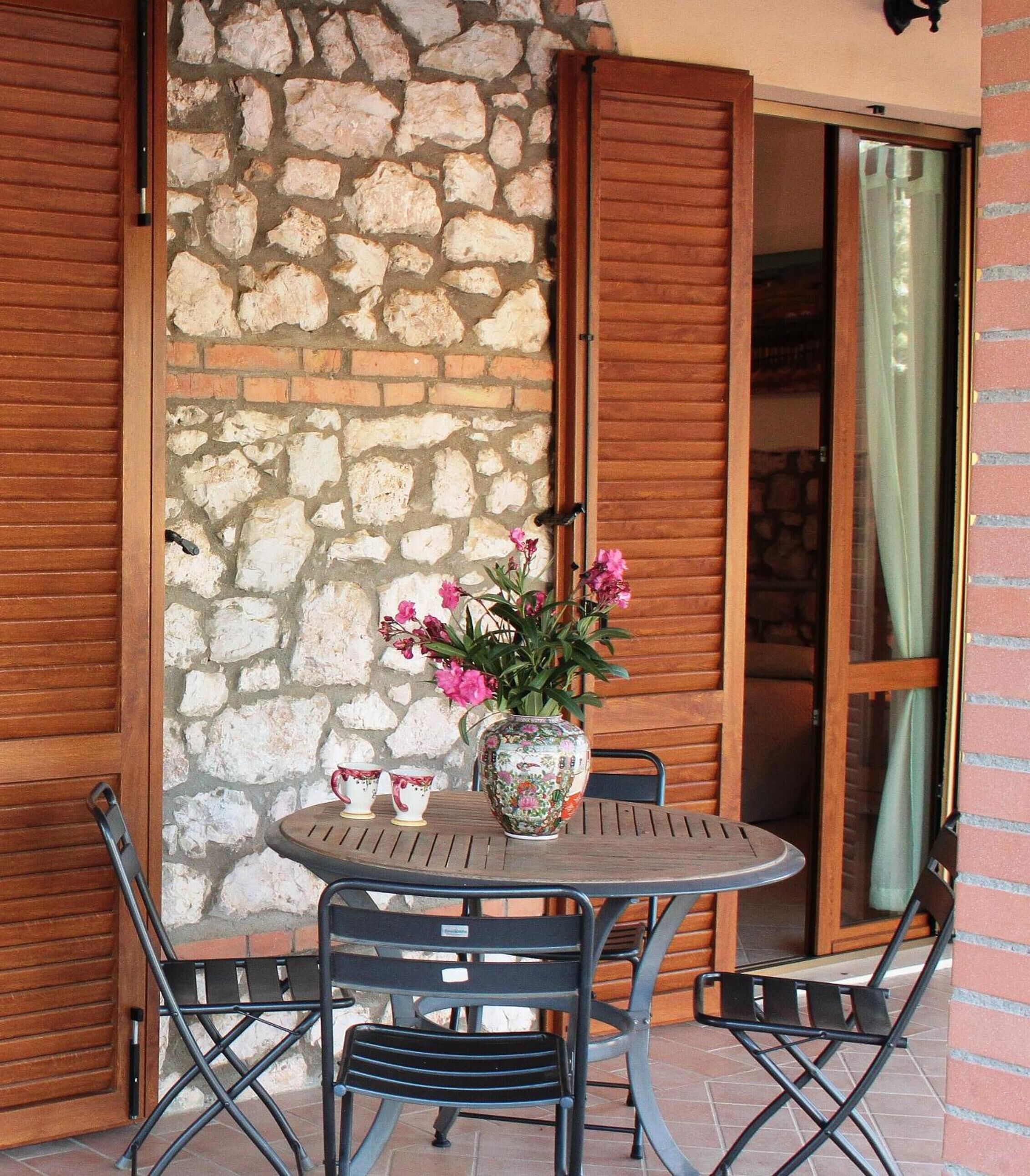 A patio with a round table, two chairs, a vase with flowers, and a stone wall.