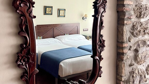 Un espejo con un marco de madera decorativo refleja una habitación con una cama, una mesita de noche y una lámpara de pared.