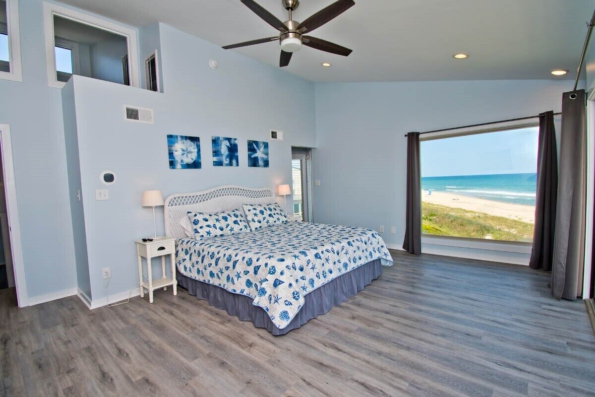 Een slaapkamer met een bed, een plafondventilator, een groot raam met uitzicht op het strand en een deur naar een andere kamer.