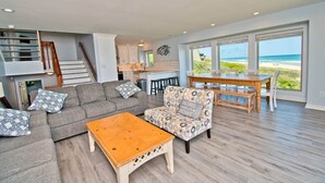 En rummelig opholdsstue med sofa, sofabord og en farverig lænestol. Et spisebord med stole står nær store vinduer med udsigt over stranden.