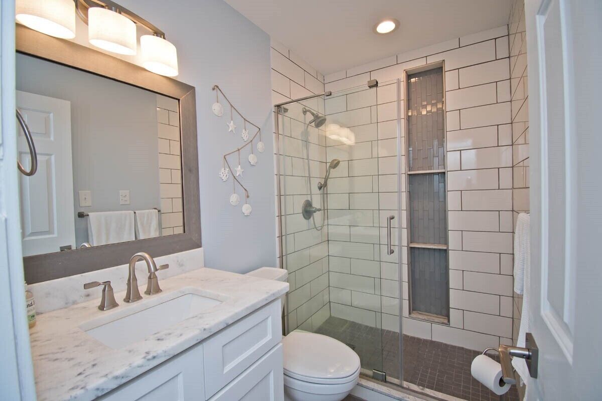 A bathroom with a white sink, mirror, and toilet, a glass-enclosed shower, and a tiled wall.