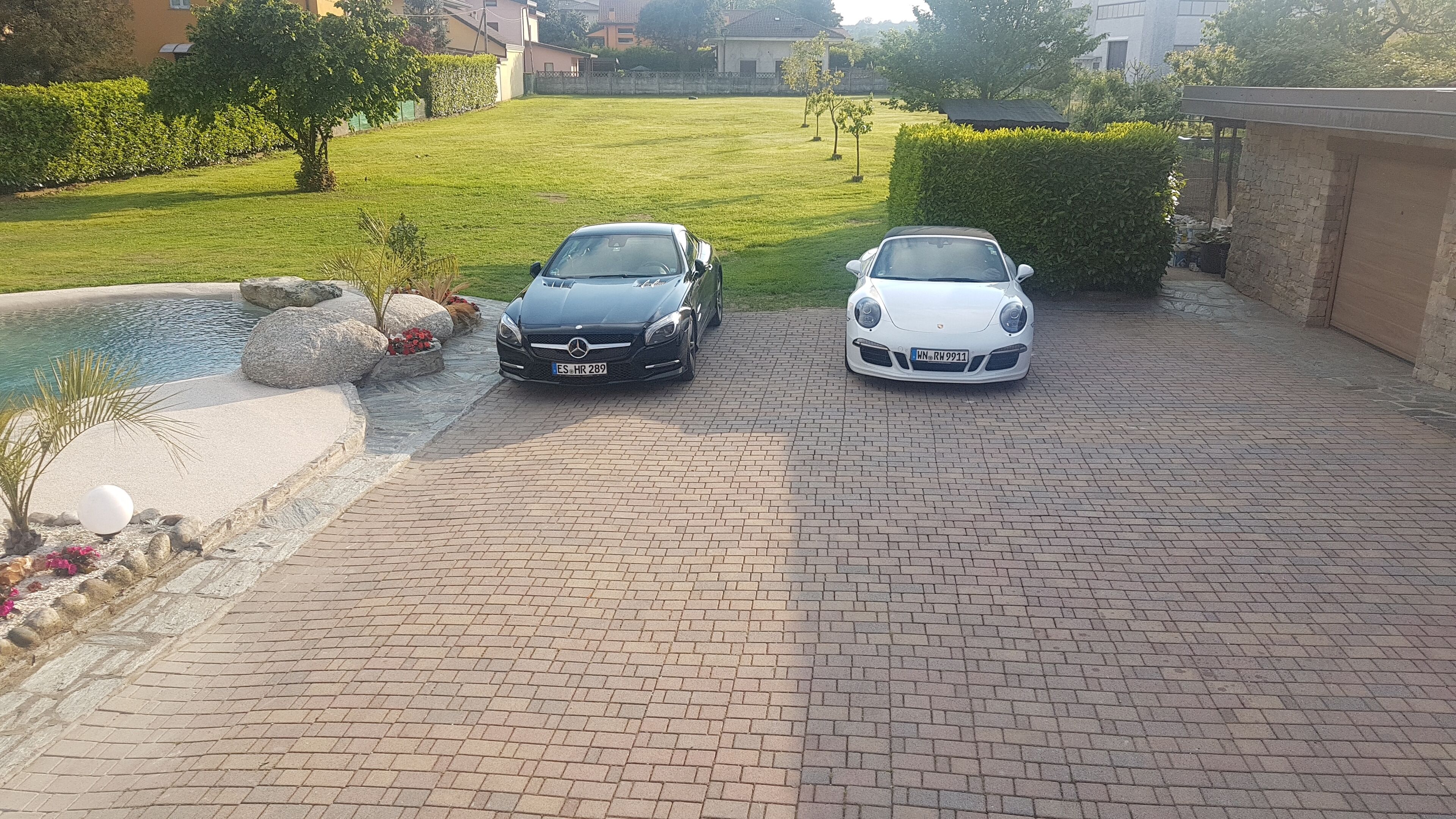 To biler parkeret i en indkørsel med brosten, en lille pool og en velplejet græsplæne.