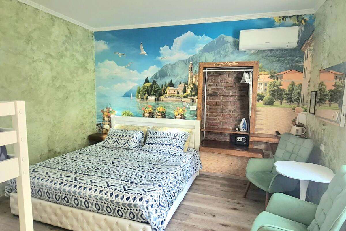 A bedroom with a bed, a chair, a small table, a wall mural, and a window.
