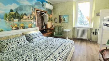 A bedroom with a bed, a green chair, a small table, a radiator, and a window with a view of trees.