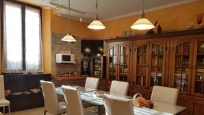 Uma área de refeições com mesa e cadeiras de vidro, uma cozinha com micro-ondas e máquina de café, e um armário de madeira com livros.