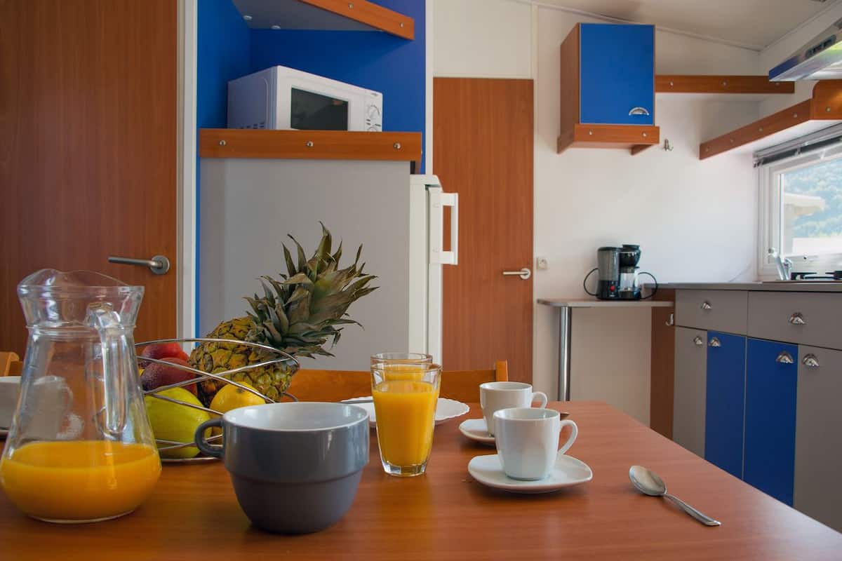 Una preparación para el desayuno que incluye un bol de fruta, una jarra de jugo de naranja y una cafetera en una cocina moderna con gabinetes azules y una ventana.