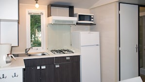 A compact kitchen with a gas stove, a microwave, and a refrigerator. There is a window providing natural light and a view of greenery outside. - Camping Le Sous Bois (Saint-Maurice-d'Ibie)