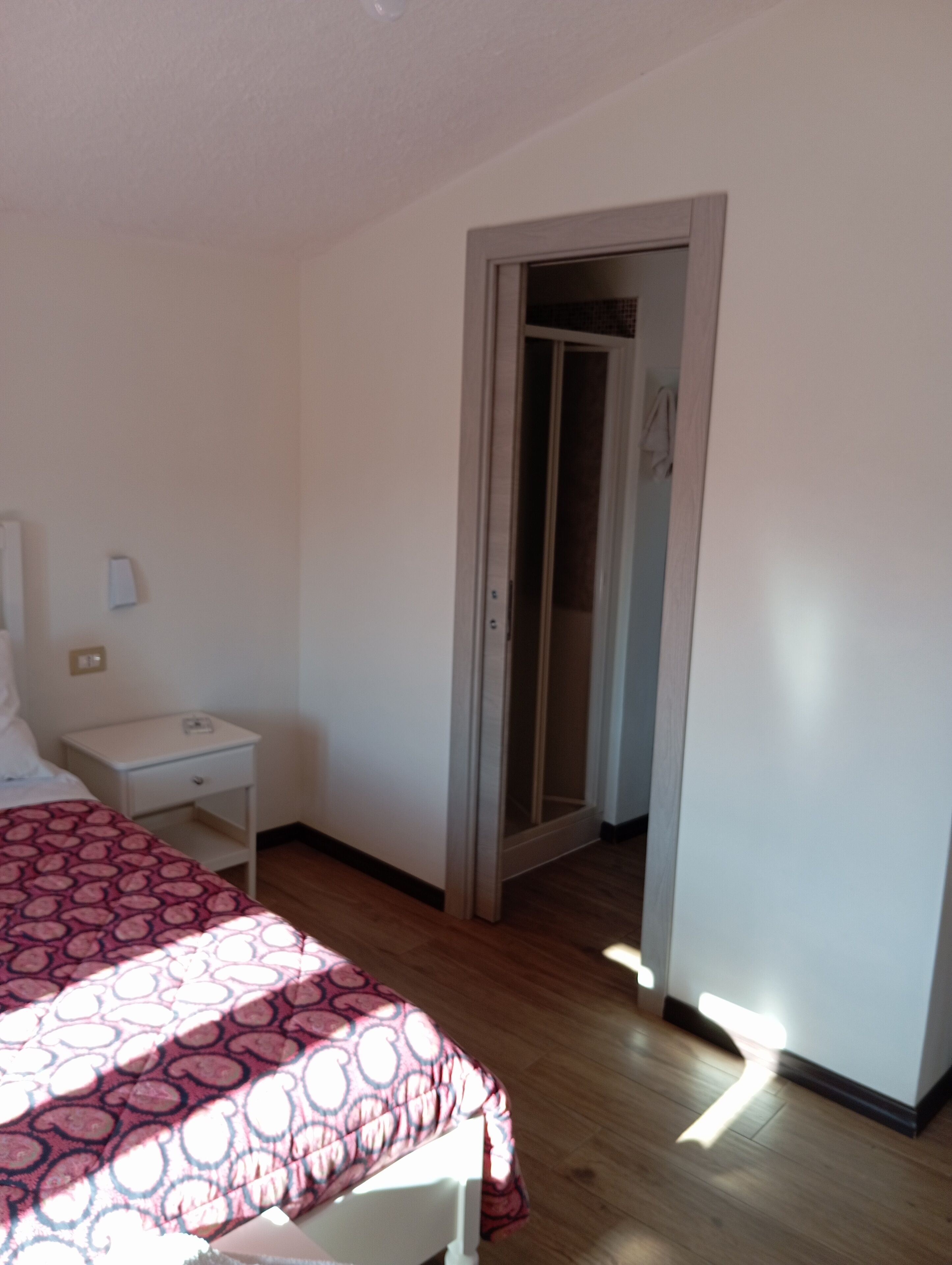 Una camera da letto con un letto, un comodino, una porta che dà su un’altra stanza e un interruttore a muro.