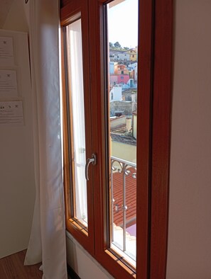 Une piĂšce avec une fenĂȘtre Ă cadre en bois, un rideau blanc et une vue sur des bĂątiments aux couleurs vives.