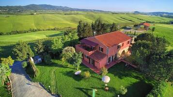 Una casa a due piani con tetto rosso, circondata da campi verdi e alberi rigogliosi.