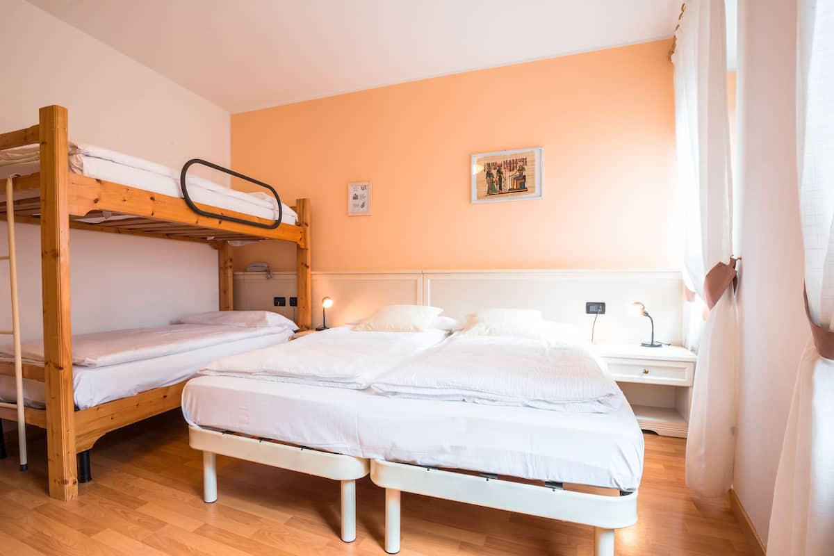 A room with two bunk beds and a single bed, wooden flooring, a nightstand with a lamp, and a wall-mounted picture.