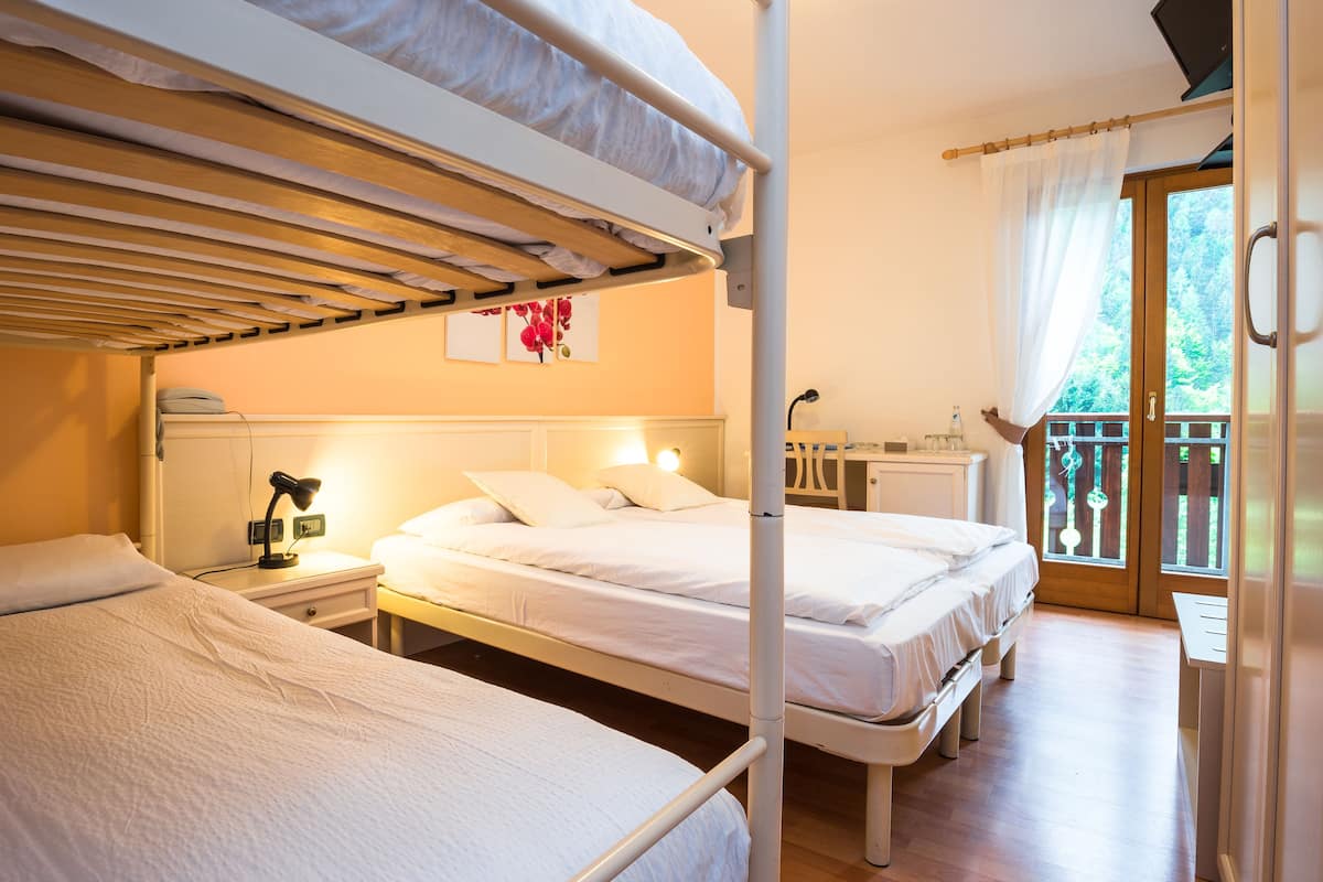 A room with two bunk beds, a desk with a lamp, a small cabinet, and a window with a view of greenery.