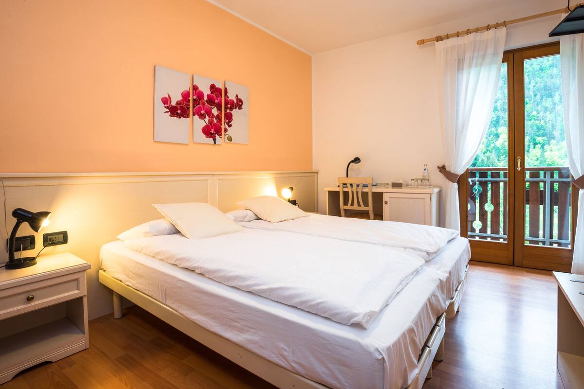 A bedroom with a bed, bedside tables, a desk, and a balcony with a view of greenery.