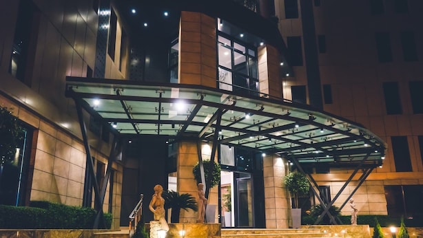 A modern hotel entrance with a glass canopy, steps leading up to the entrance, and two statues on the steps. - Belanova Apartments & Suites (Abuja)