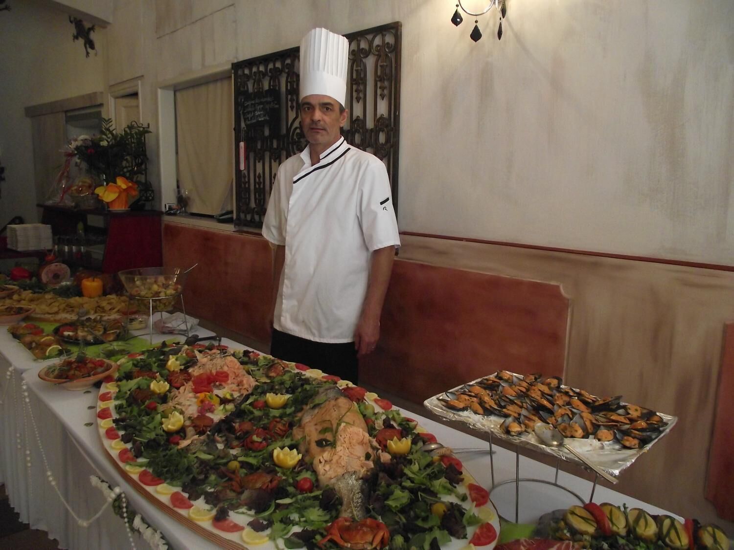 A buffet table with various dishes, a chef in uniform, and a decorative wall.