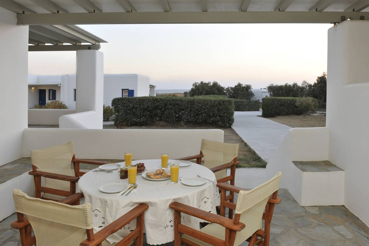 A covered outdoor dining area with a table set for two, surrounded by wicker chairs, overlooking a landscaped garden with white buildings in the background.