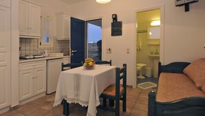 A compact kitchen with white cabinets, a dining table with a white tablecloth, and a fruit bowl. There is a blue door leading to a bathroom, and a TV mounted on the wall. - Golden Sand Paros (Paros)