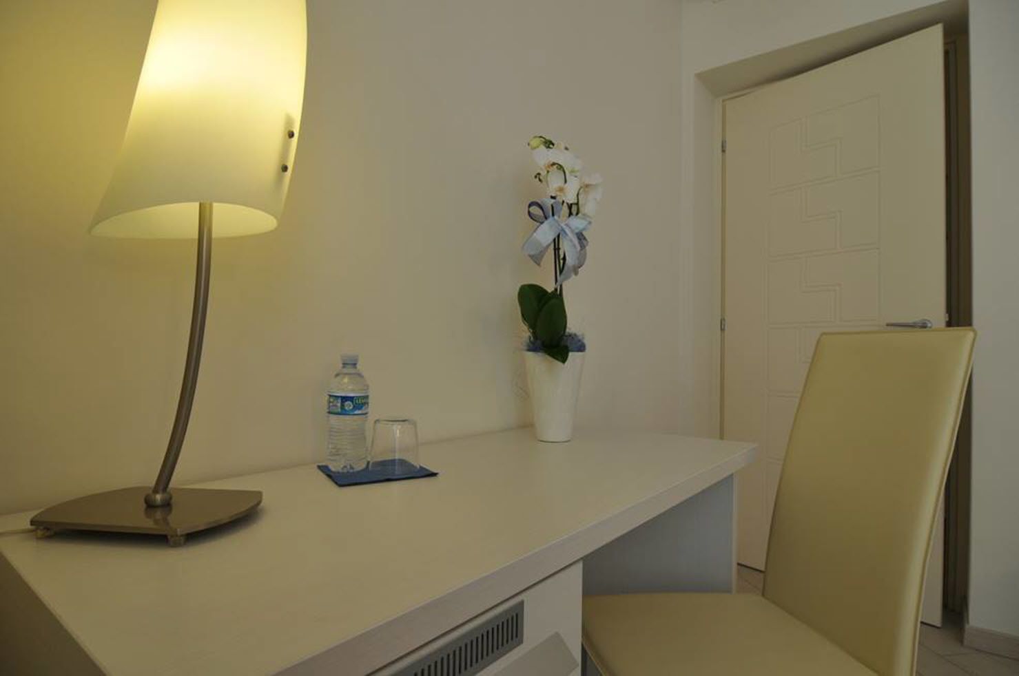 A hotel room with a desk, a lamp, a water bottle, a glass, and a potted plant.