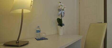A hotel room with a desk, a lamp, a water bottle, a glass, and a potted plant.