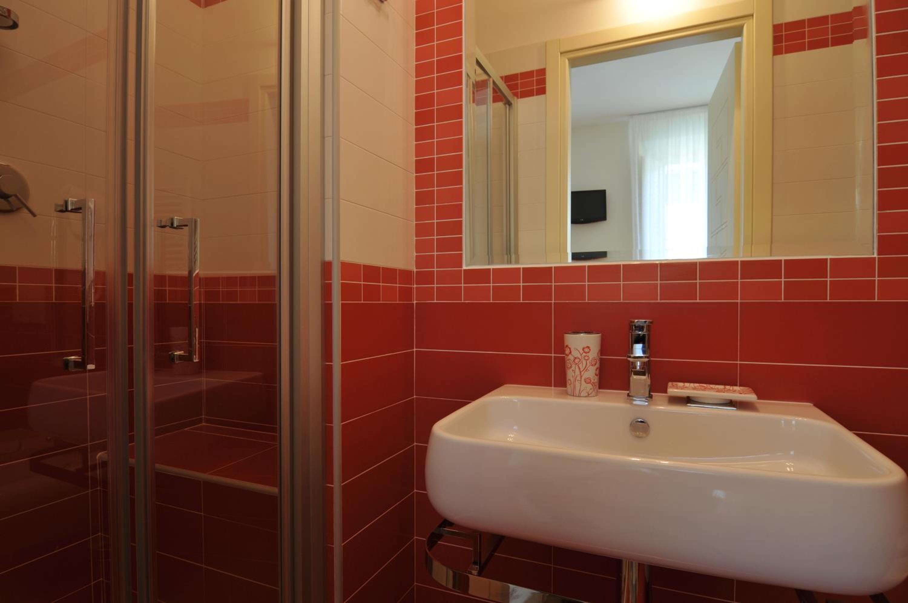 Ein Badezimmer mit Duschkabine aus Glas, einem weißen Waschbecken mit Spiegel darüber und roten Fliesenwänden.