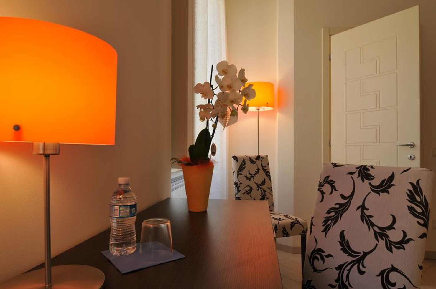 Une chambre d’hôtel avec une table en bois, deux chaises, une bouteille d’eau, un verre et une orchidée en pot.
