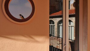 Una ventana circular con un pájaro afuera, un balcón con barandilla de metal, y un edificio con ventanas de arco y tejado de tejas.