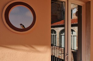 Una ventana circular con un pájaro afuera, un balcón con barandilla de metal, y un edificio con ventanas de arco y tejado de tejas.