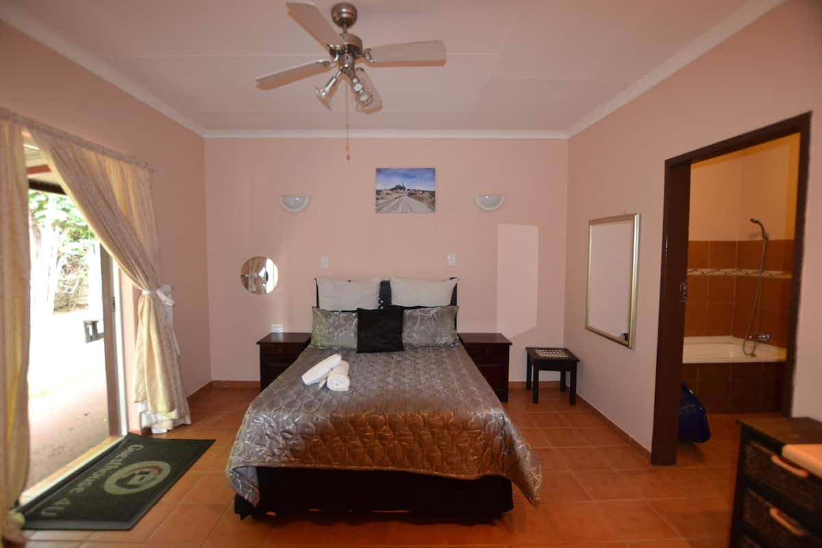 A bedroom with a bed, a ceiling fan, a nightstand, a small table, a door leading to a bathroom, and a view of the outside.