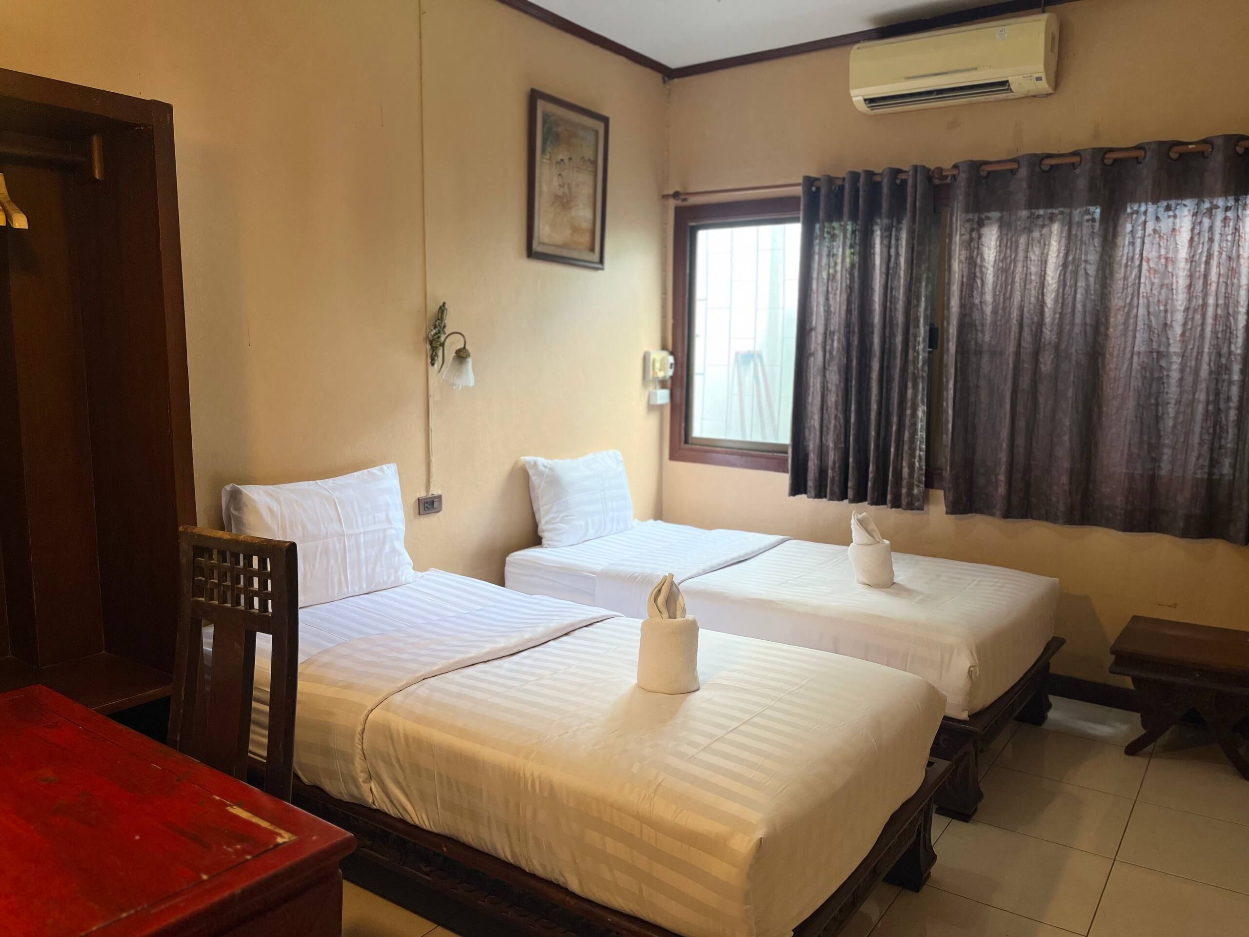 A hotel room with two beds, a wooden chair, a small table, a window with curtains, and an air conditioning unit.