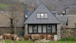 Uma casa com fachada de pedra e telhado escuro, cercada por um campo gramado com vacas.